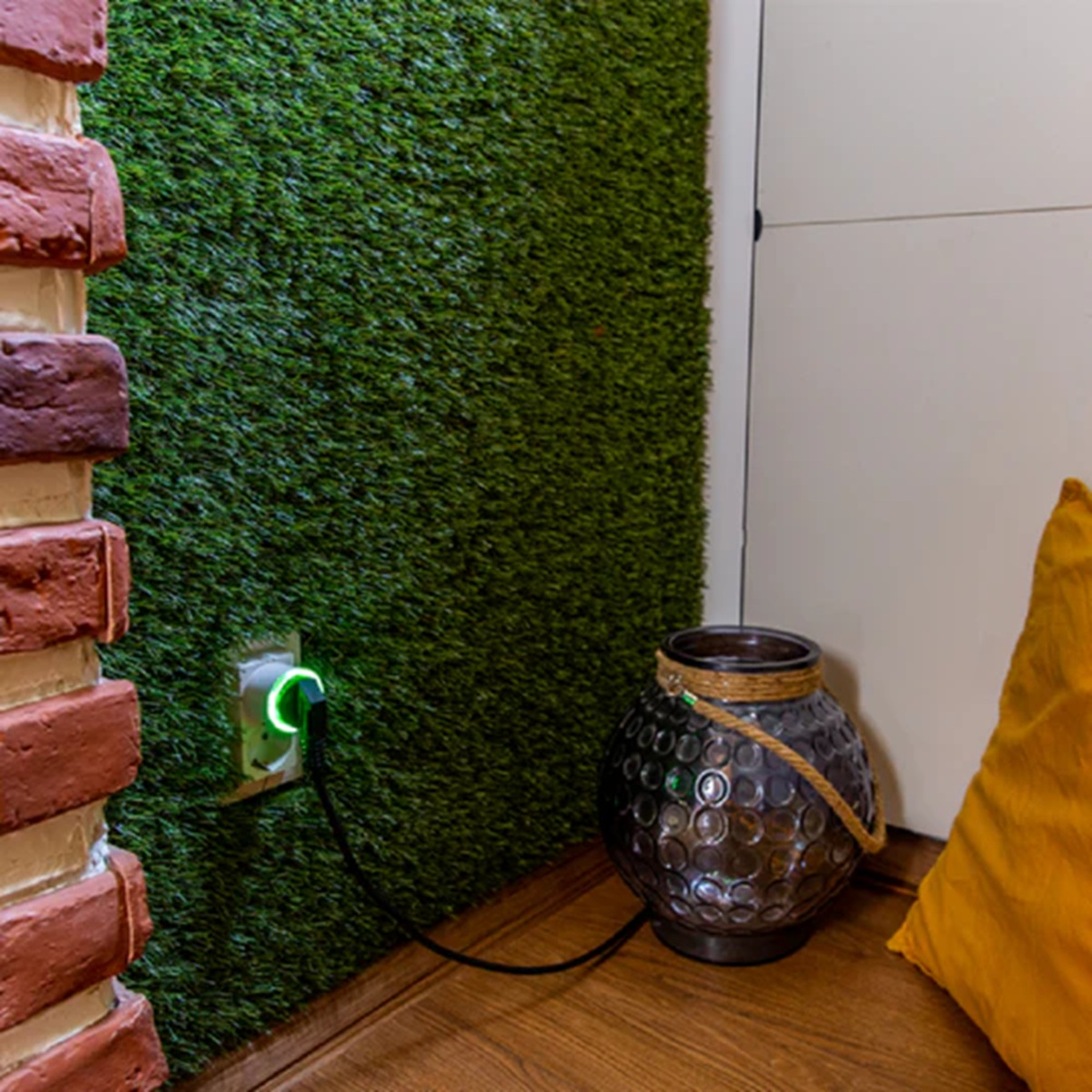 Ściana z zieloną sztuczną trawą i gniazdem elektrycznym. Obecny jest dekoracyjny wazon i poduszka.