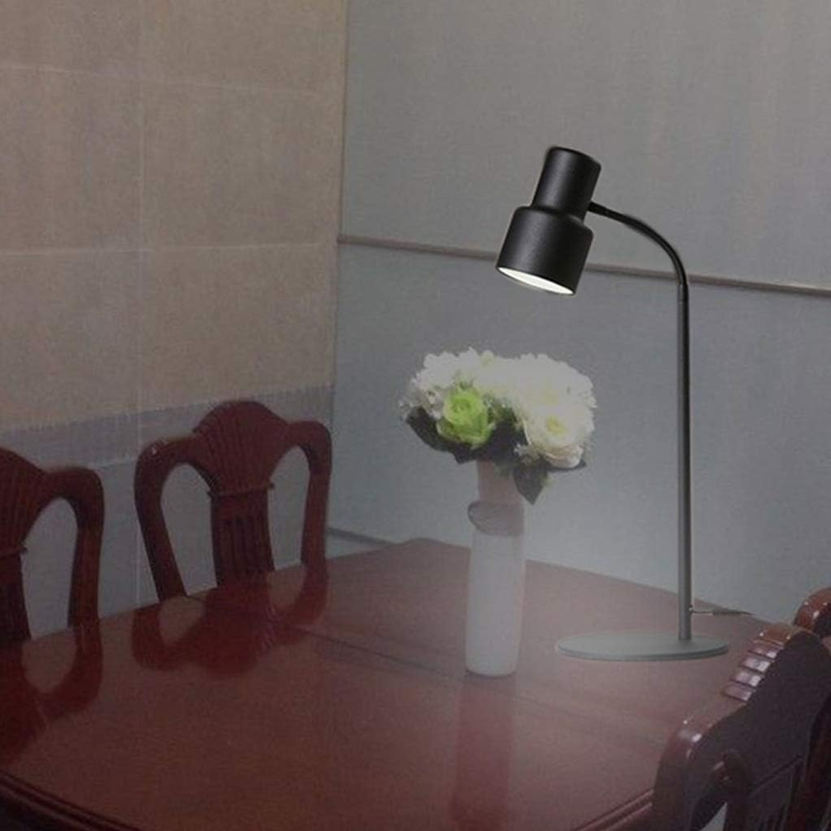 Czarna lampa biurkowa oświetla stół. Na stole wazon z kwiatami, w tle krzesła.