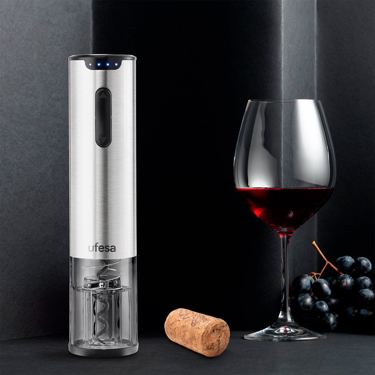 Abridor de vino eléctrico plateado con copa y vino tinto, un corcho y uvas sobre una superficie oscura.