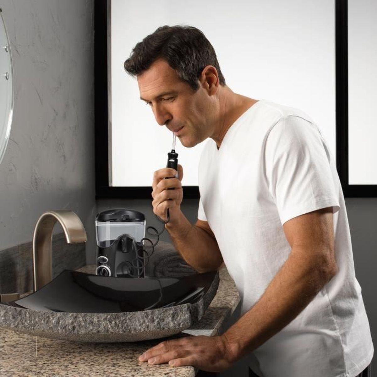Mężczyzna używający irygatora w łazience, pochylony nad umywalką. Trzyma irygator w ustach.