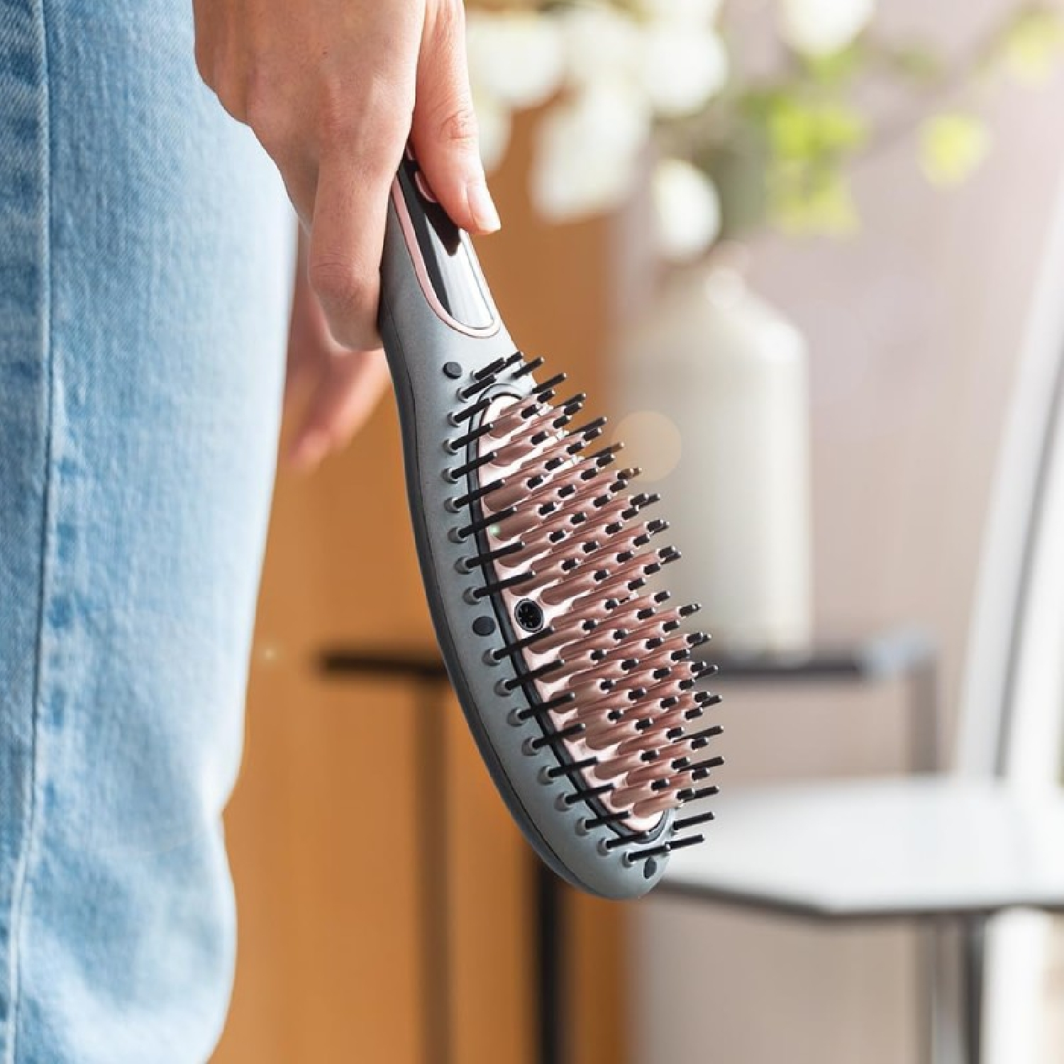 Una persona tiene in mano una spazzola grigia per capelli con setole rosa, vicino a una finestra e uno sfondo sfocato.