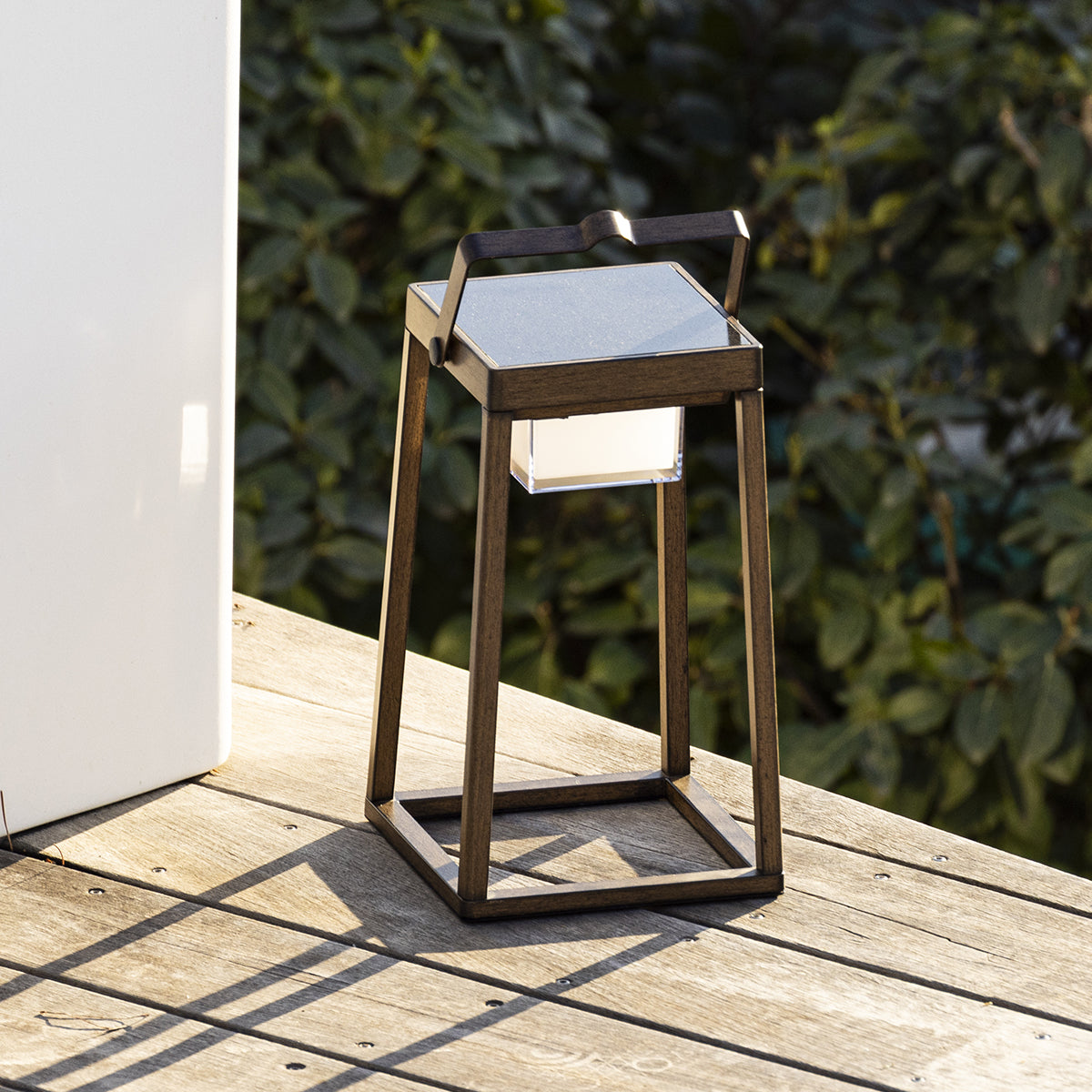 Laterne im Freien auf einer Holzterrasse, mit einer weißen Struktur im Hintergrund und Sonnenlicht.