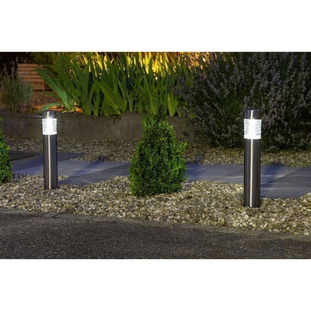 Zwei silberne zylindrische Lichter in einem Garten. Kies und Büsche im Hintergrund.