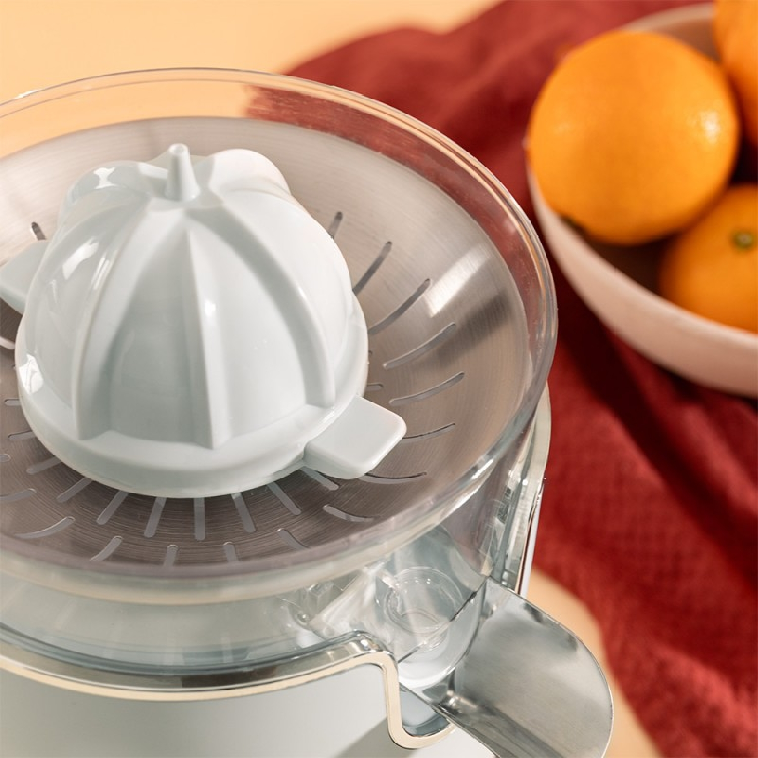 Exprimidor de cítricos, plástico blanco y transparente. Naranjas y tela roja al fondo.