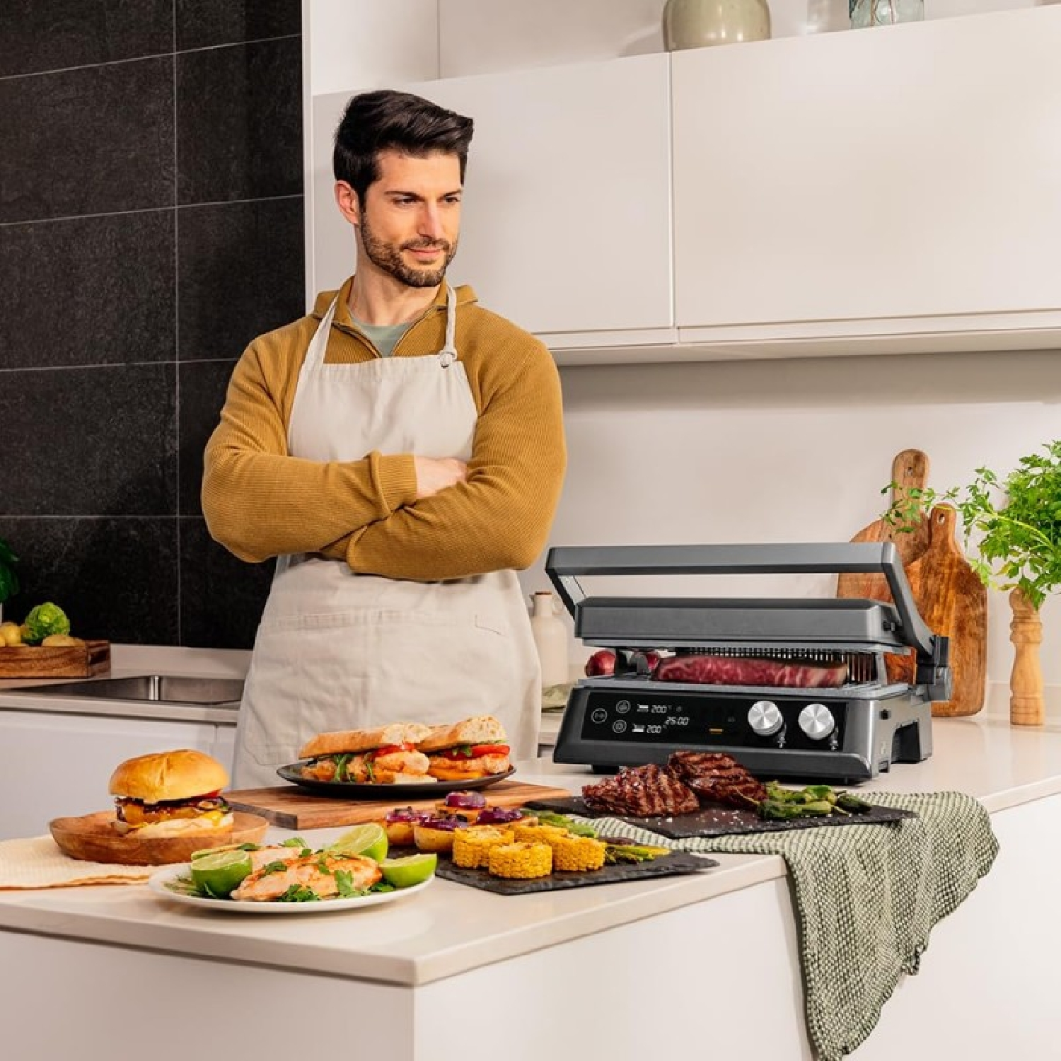Ein Mann mit Schürze grillt Essen auf einem Grill. Auf einer Theke stehen Teller mit Essen. Der Hintergrund ist eine Küche.