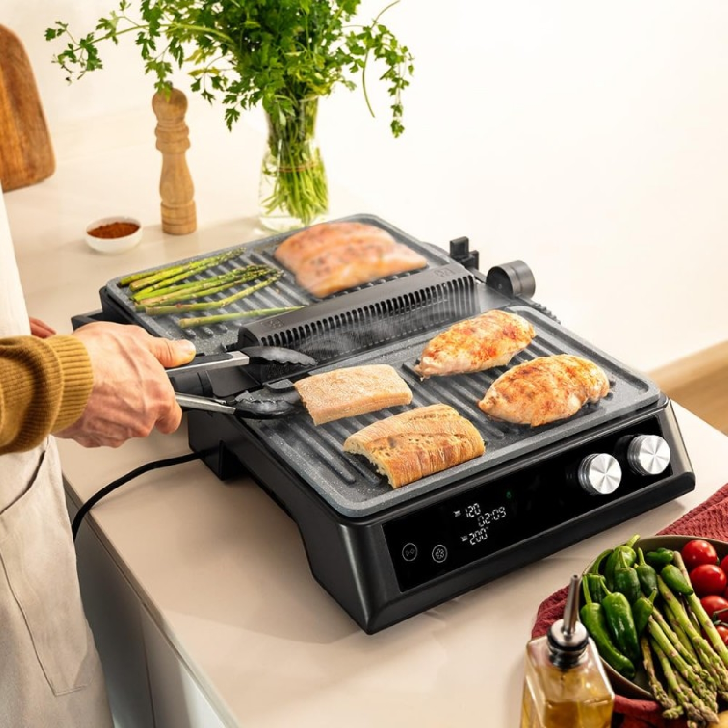 Ein elektrischer Grill mit Hühnchen, Spargel und Brot. Eine Person greift mit einer Zange nach Essen. Gemüse und Gewürze sind in der Nähe.