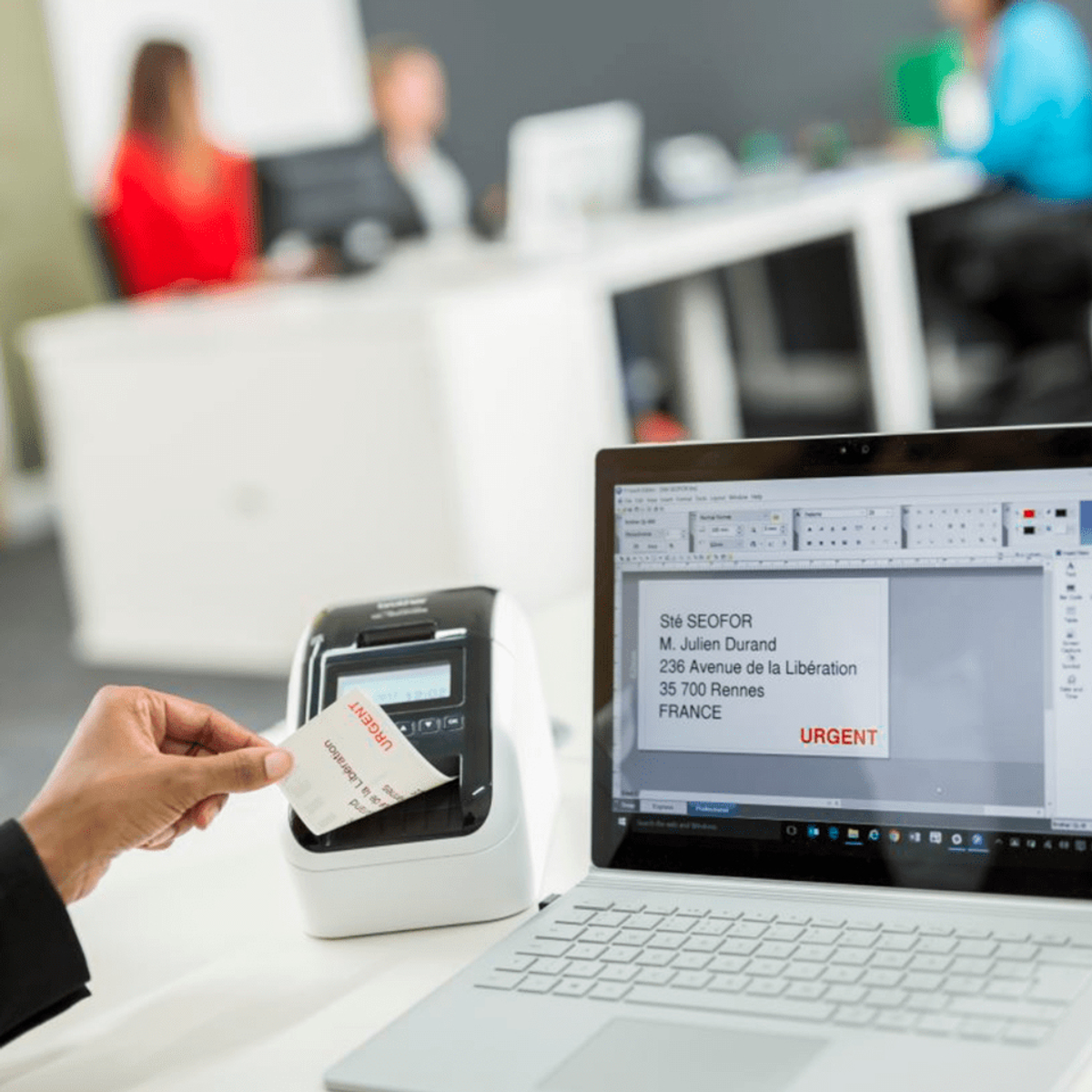 Eine Person hält ein Etikett in der Nähe eines Etikettendruckers. Ein Laptop und Personen sind im Hintergrund.