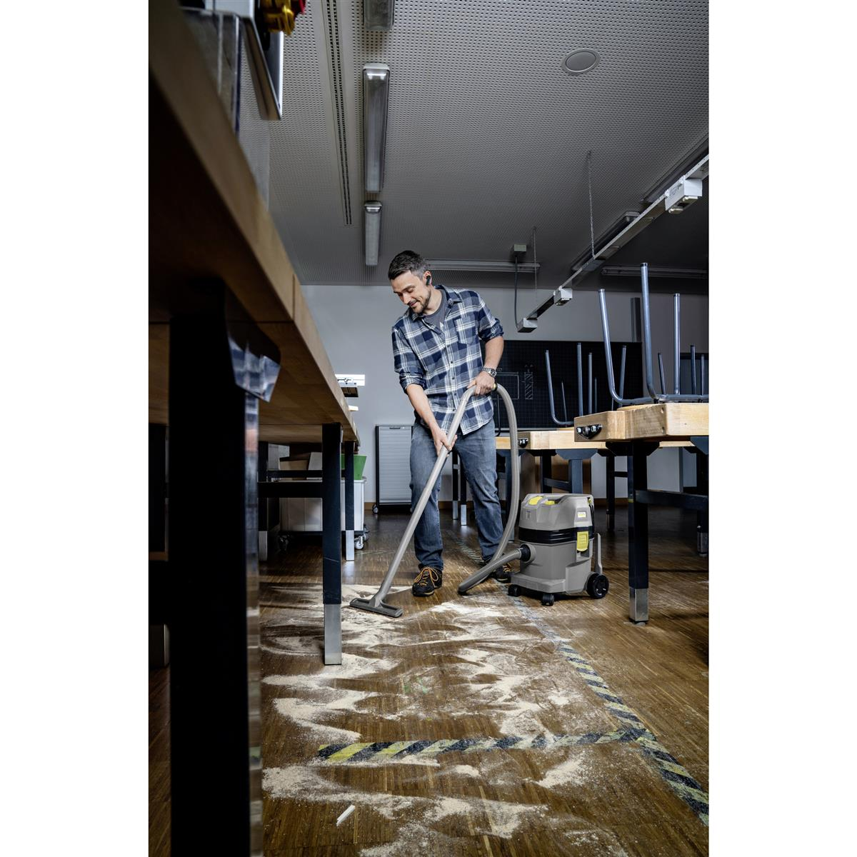 Uomo che aspira il pavimento in legno in un laboratorio. Un aspirapolvere grigio e tavoli sullo sfondo.