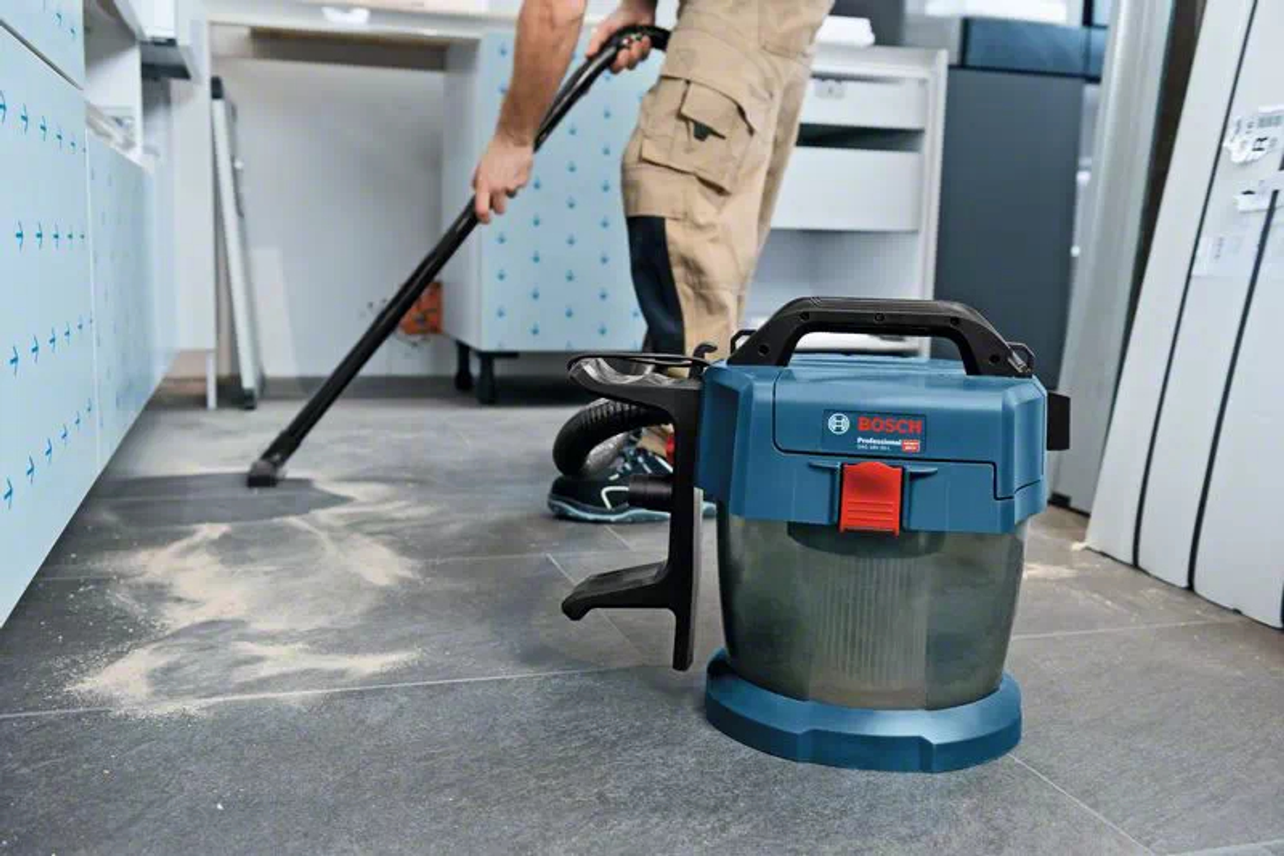 Una persona aspira un pavimento con un aspirapolvere blu e nero, con polvere visibile sul pavimento.
