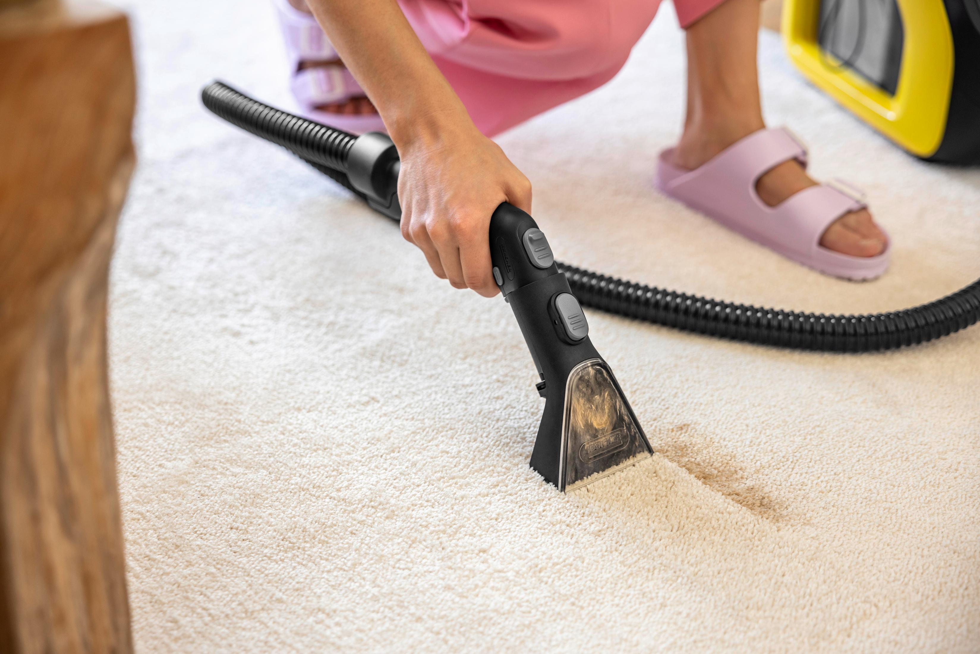 Personne aspirant un tapis blanc avec un aspirateur noir. Des sandales roses sont visibles aux pieds.