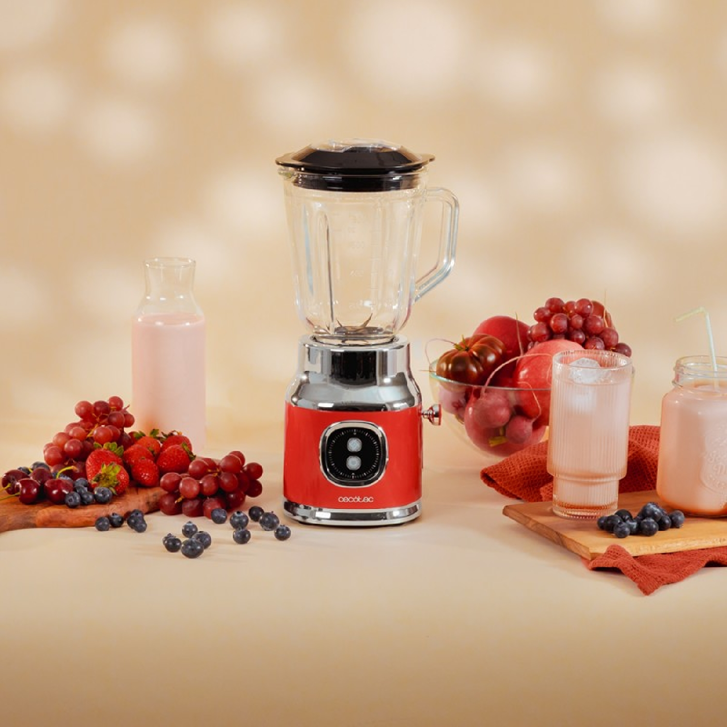 Frullatore con base rossa, caraffa in vetro e vari frutti su superficie beige.