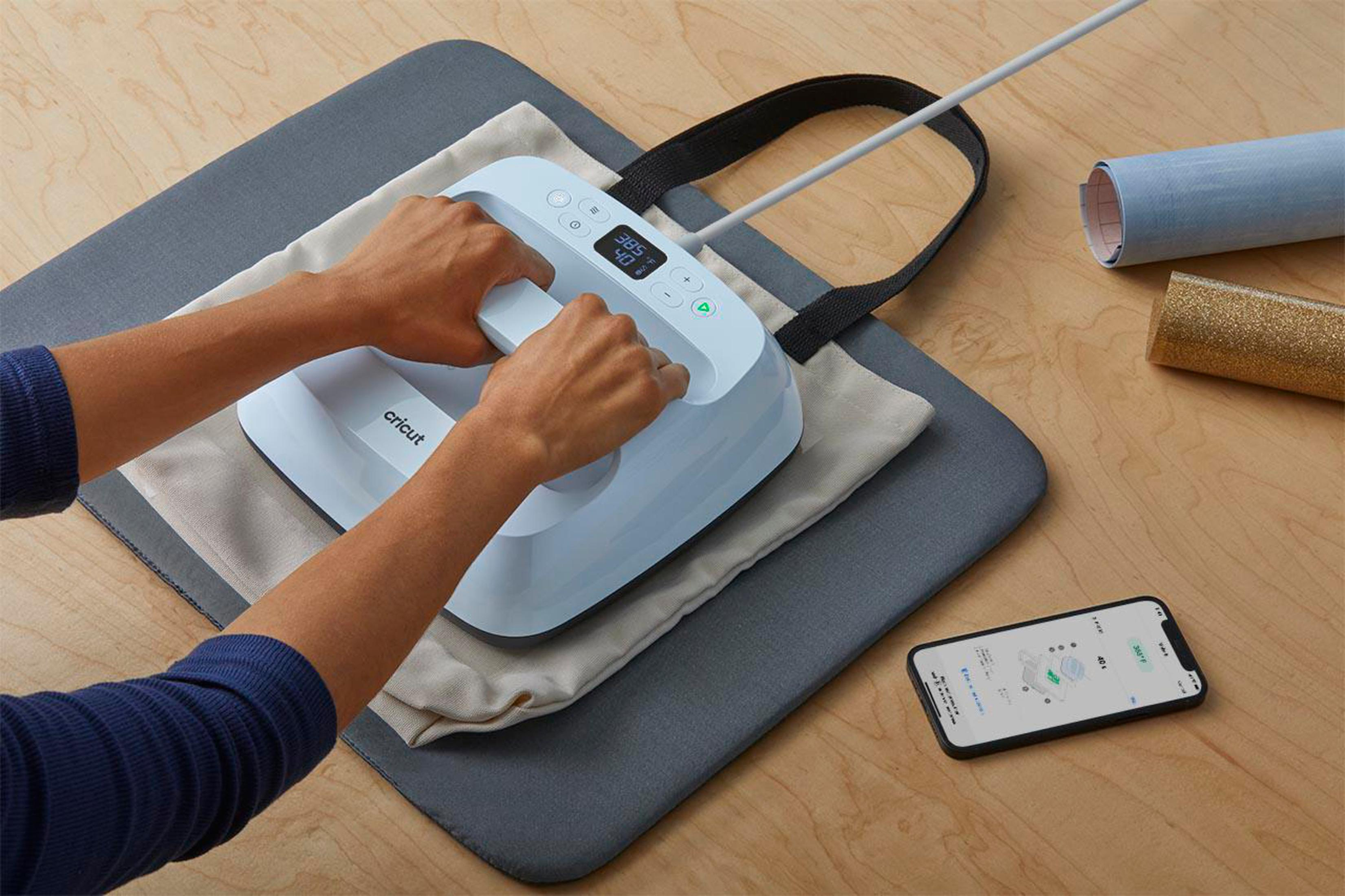 Person verwendet eine Cricut EasyPress auf einer Tragetasche. Ein Telefon zeigt eine App mit Designdetails.