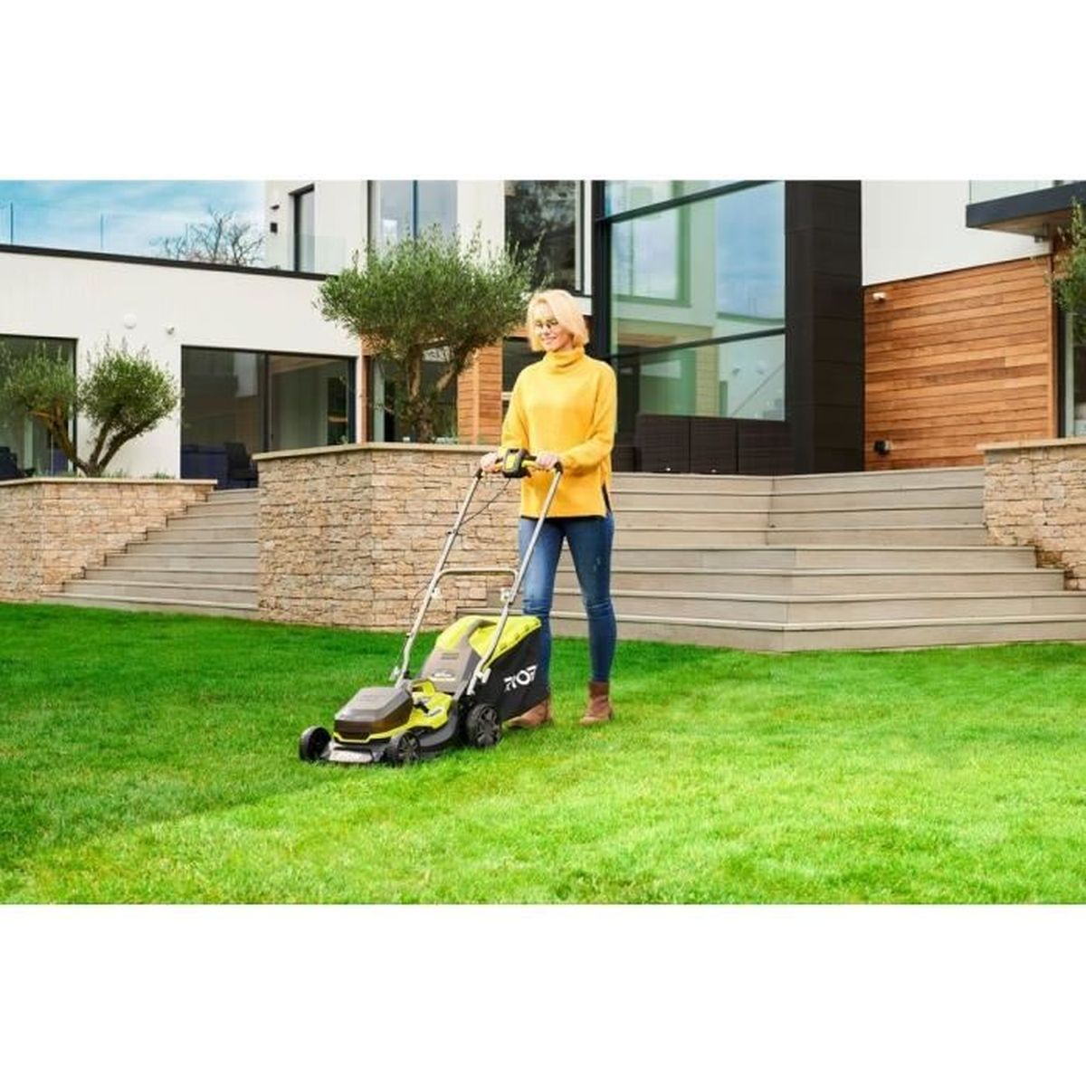 Frau mäht Rasen mit Ryobi Rasenmäher. Sie trägt Gelb und Blau. Haus im Hintergrund.