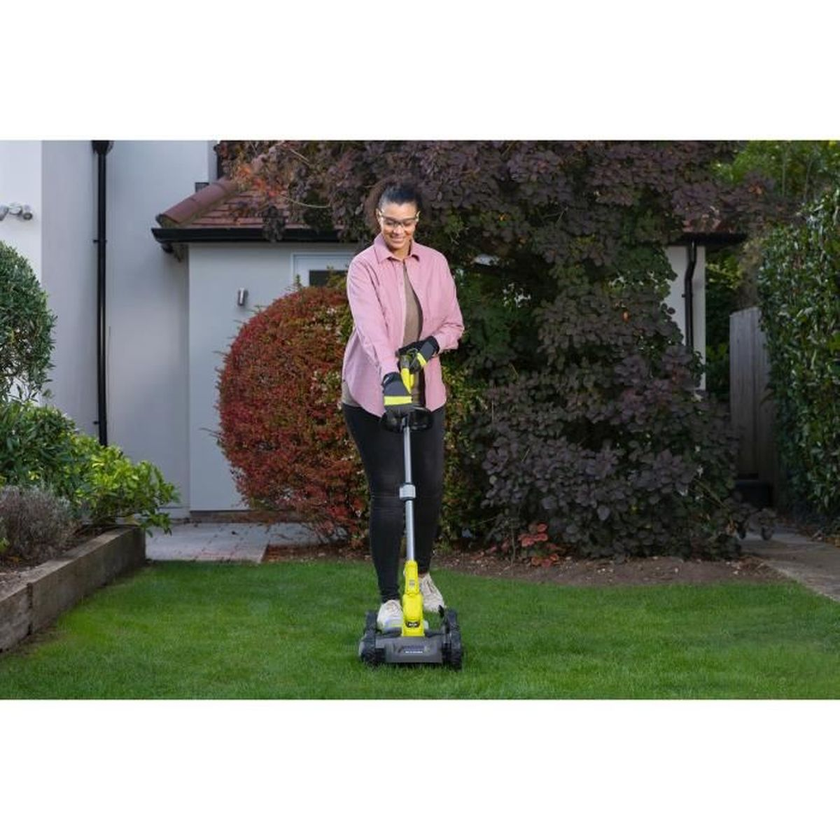 Eine Frau in einem rosa Hemd benutzt einen Rasenkantenschneider auf grünem Gras. Ein Haus und Bäume sind im Hintergrund.