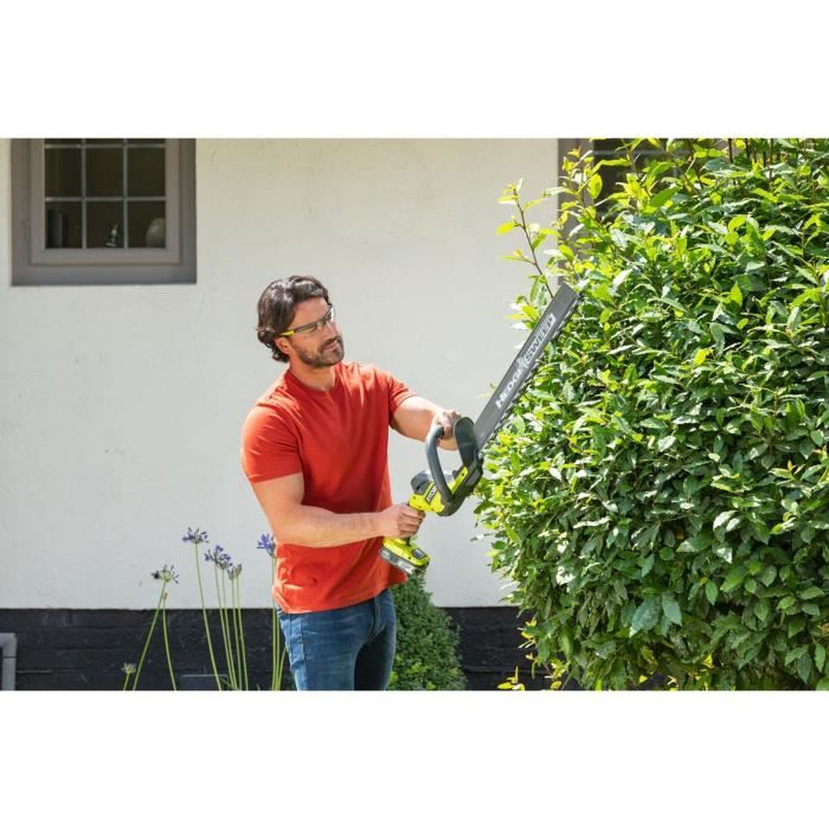 Ein Mann benutzt eine Heckenschere an einem Busch, trägt eine Brille, in einem Garten an einem sonnigen Tag.