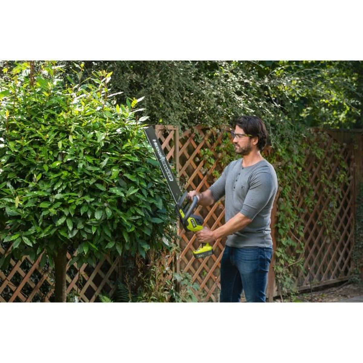 Mann trimmt eine Hecke mit einem Elektrowerkzeug. Grüne Hecke und Holzzaun.