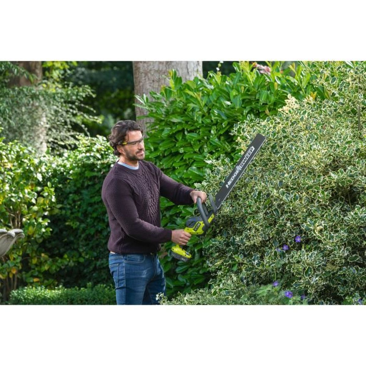 Ein Mann mit Brille schneidet eine Hecke mit einem elektrischen Heckenschneider. Grüne Hecke rechts.