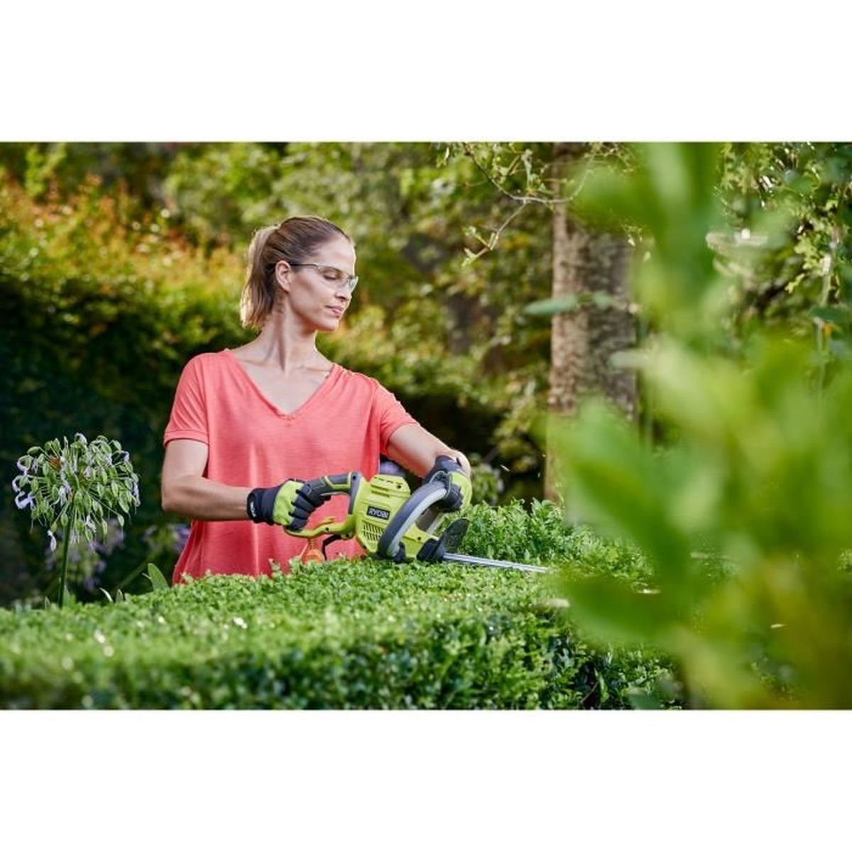 Frau im rosa Hemd schneidet eine grüne Hecke mit einer elektrischen Heckenschere.