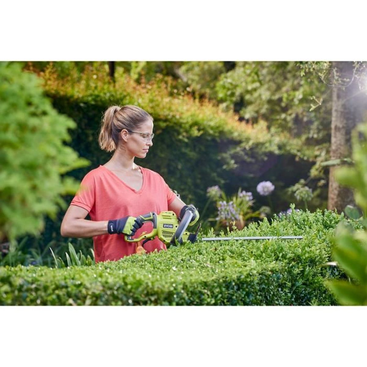 Eine Frau mit Schutzbrille schneidet eine grüne Hecke mit einer elektrischen Heckenschere in einem Garten.