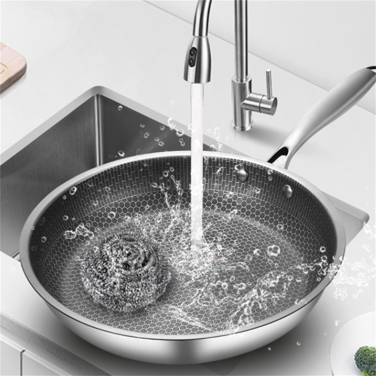 Edelstahlpfanne in einem Waschbecken, wird mit Wasser gewaschen. Ein Schrubber ist in der Pfanne.