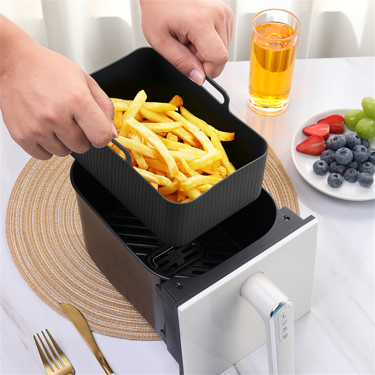 Hände, die einen schwarzen Behälter mit Pommes aus einer Heißluftfritteuse nehmen. Obst und Saft auf dem Tisch.
