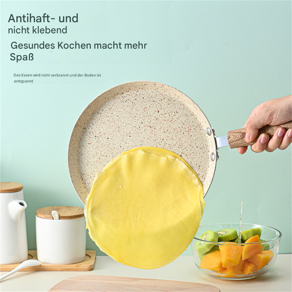 Eine Pfanne mit Pfannkuchen. Eine Hand hält die Pfanne mit Holzgriff. Zutaten liegen daneben.