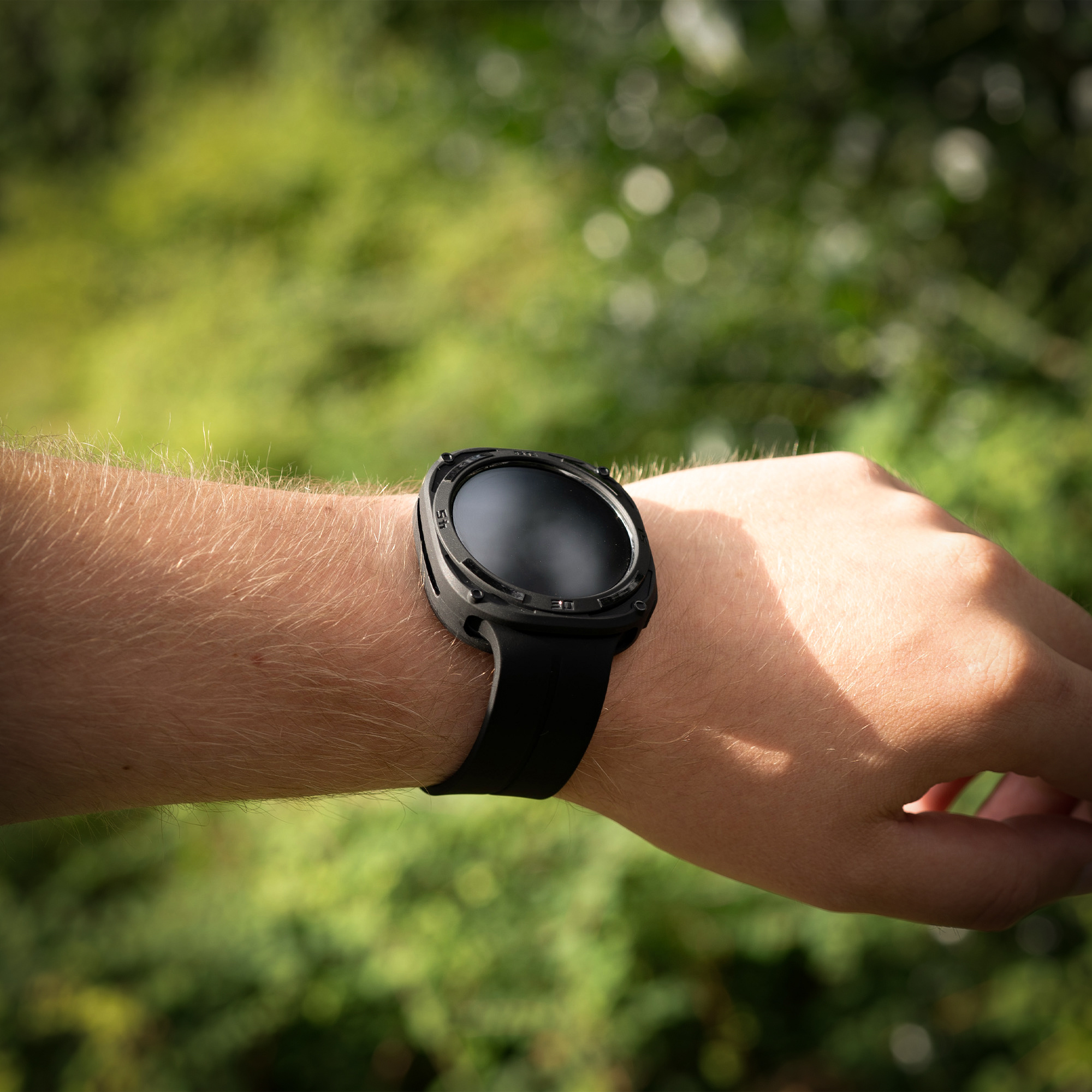 Handgelenk mit schwarzer Smartwatch mit schwarzem Armband. Verschwommenes grünes Laub im Hintergrund.