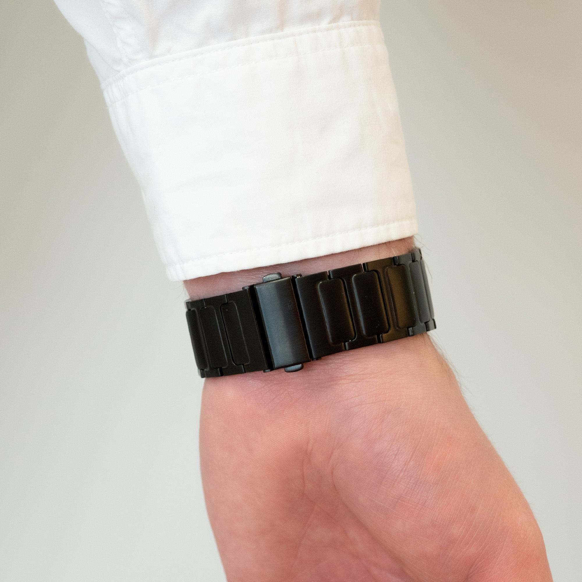 Ein schwarzes Uhrenarmband am Handgelenk, ein weißes Hemd im Hintergrund.