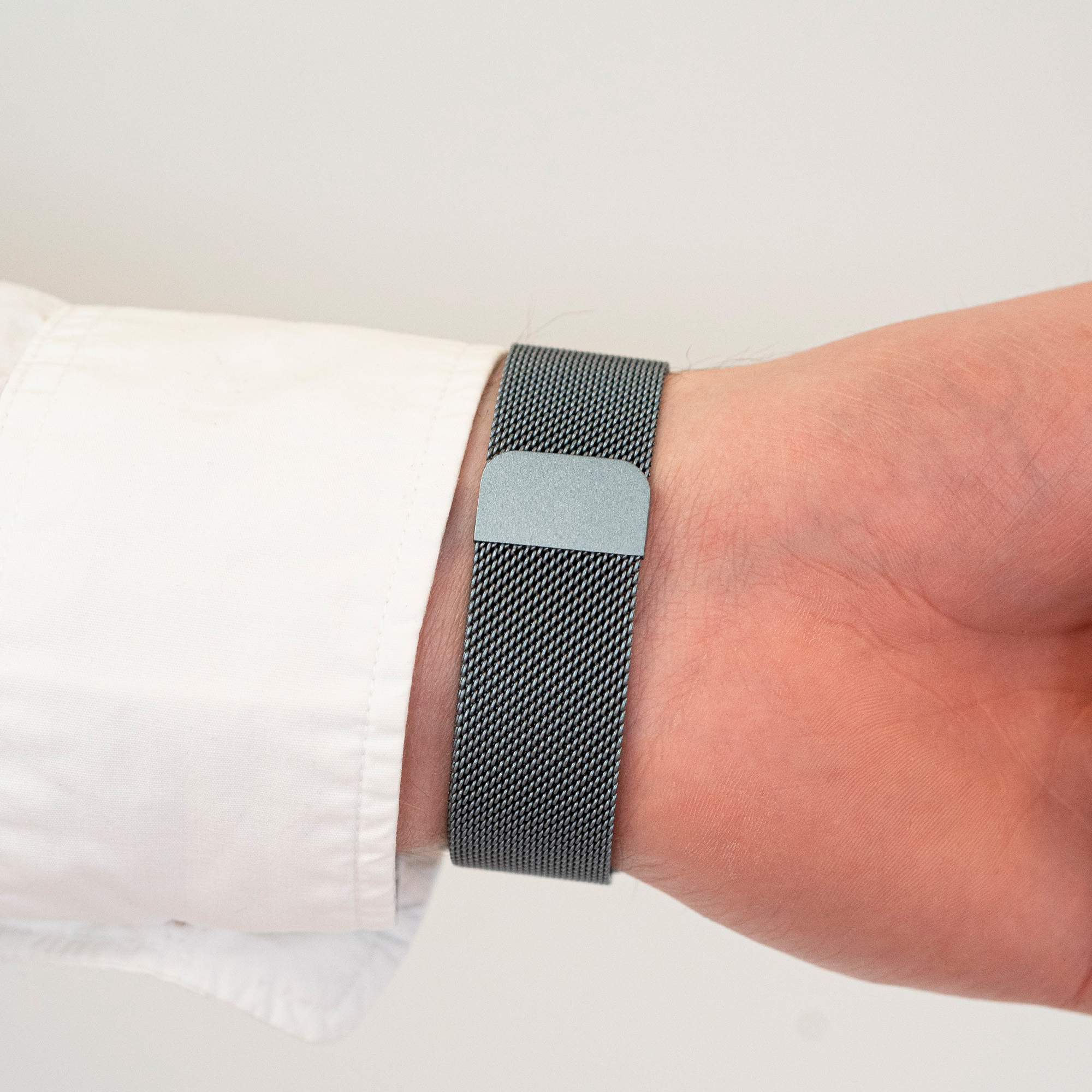 Das Handgelenk einer Person mit einem dunkelgrauen Mesh-Armband und einem weißen Hemdärmel.
