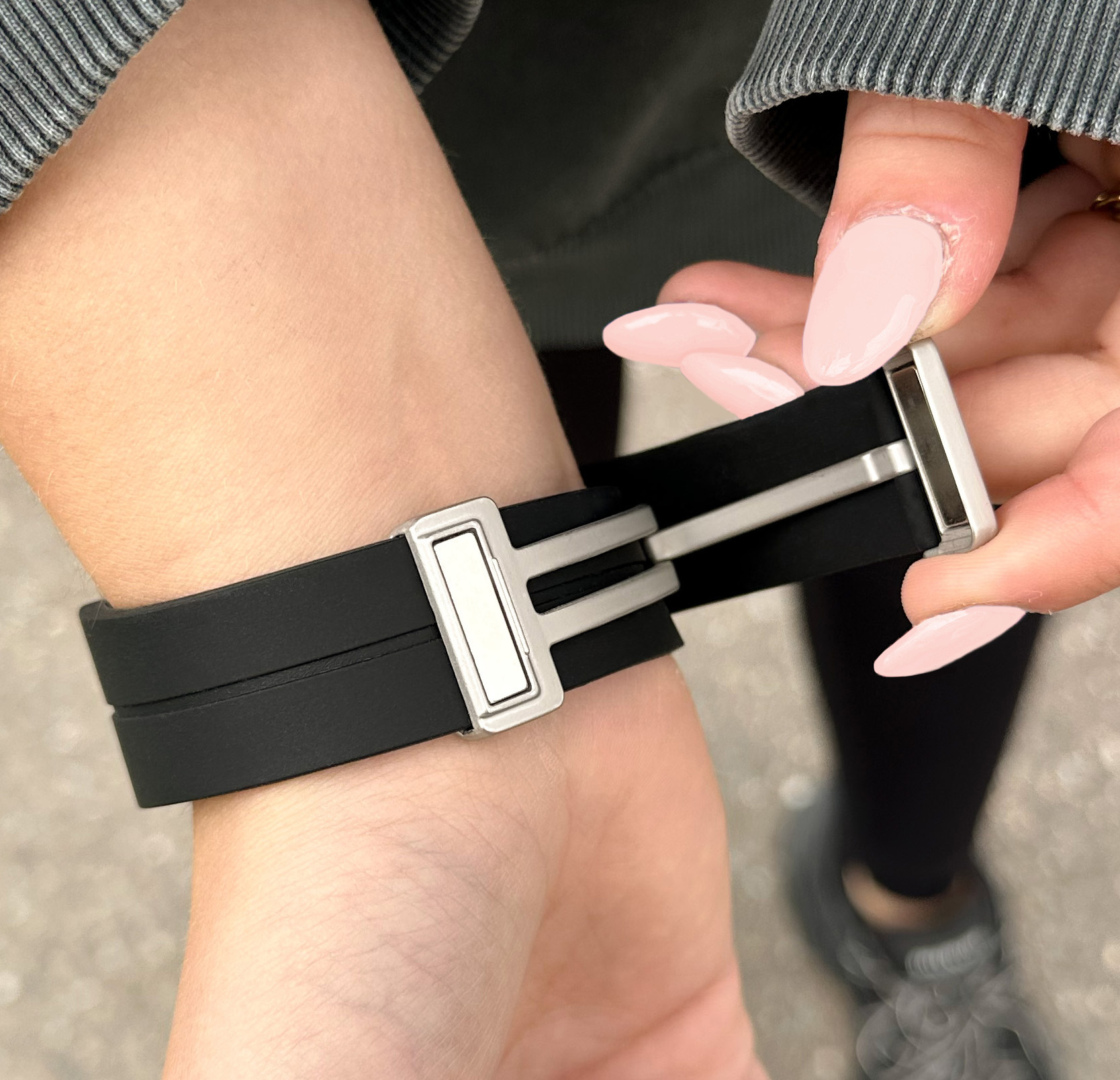 Die Hand einer Person befestigt ein schwarzes Uhrenarmband mit silberner Schließe am Handgelenk. Rosa Nagellack ist sichtbar.
