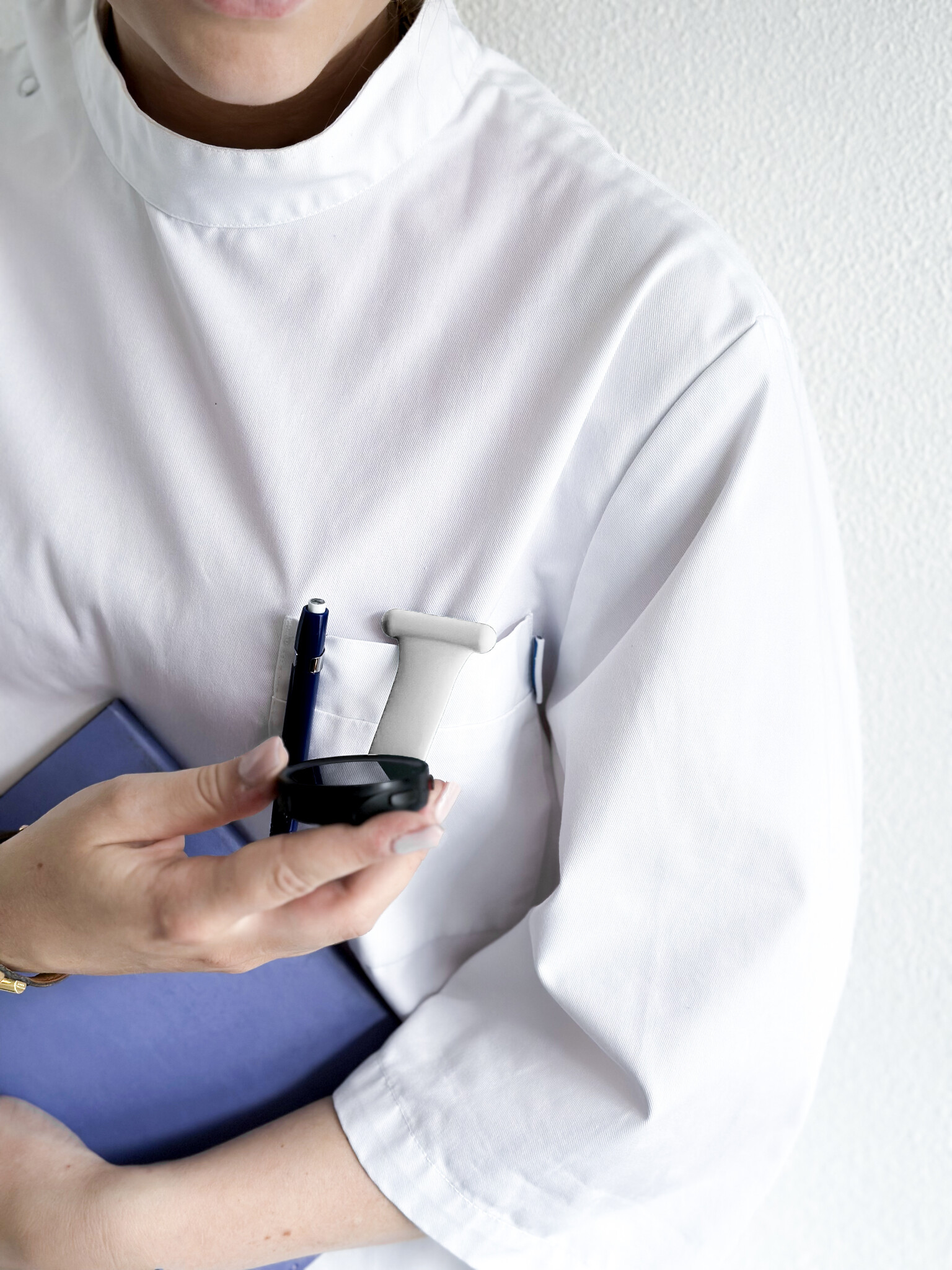 Eine Person in weißem Kittel hält einen blauen Ordner und einen Stift. Eine schwarze Uhr ist in ihrer Tasche.