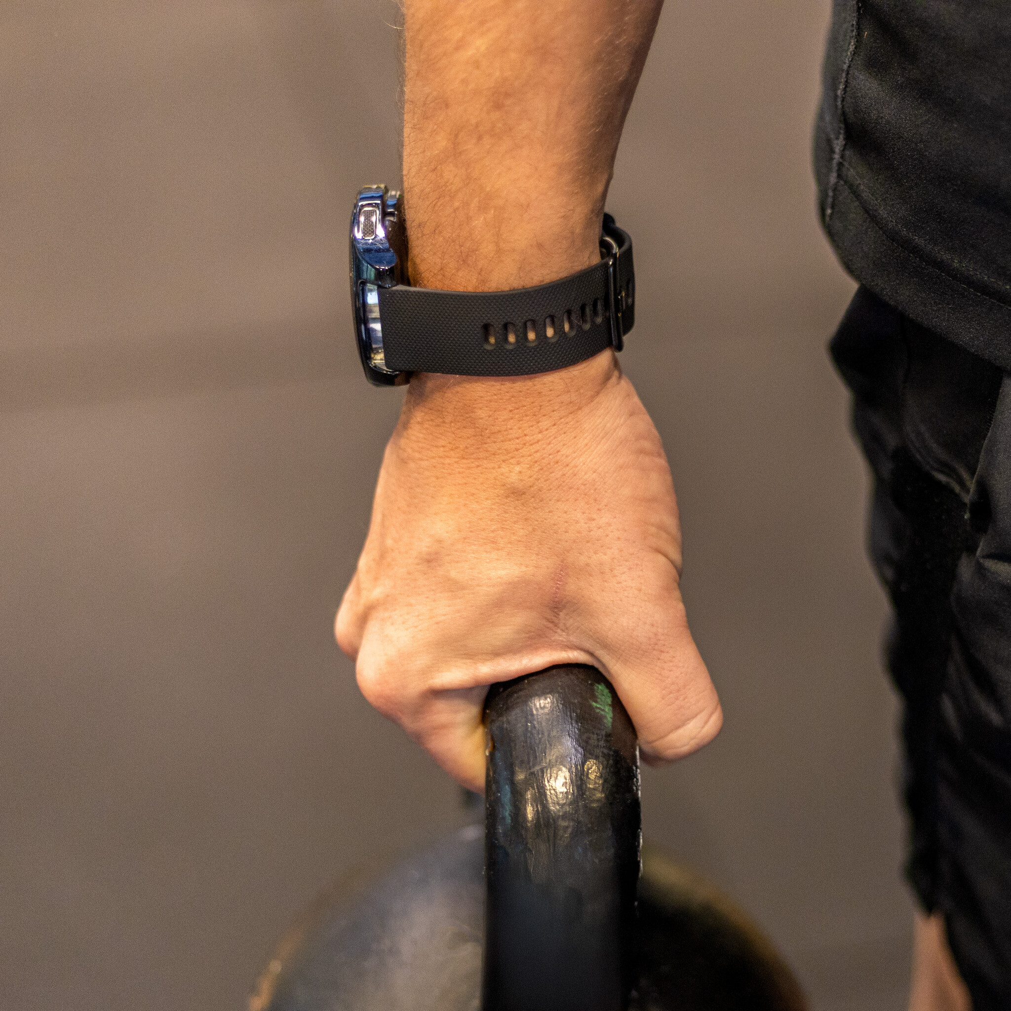 Die Hand einer Person hält eine Kugelhantel, trägt eine schwarze Uhr mit schwarzem Armband. Gymnastikhalle.