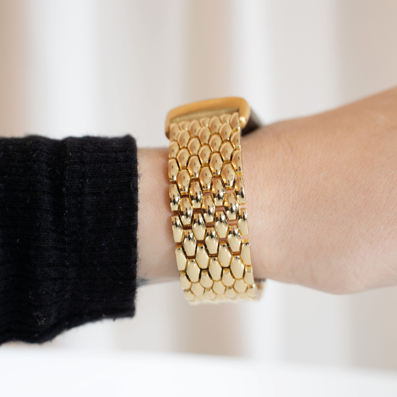 Handgelenk einer Person mit einem goldenen Uhrenarmband. Das Armband besteht aus kleinen, verbundenen Goldformen.