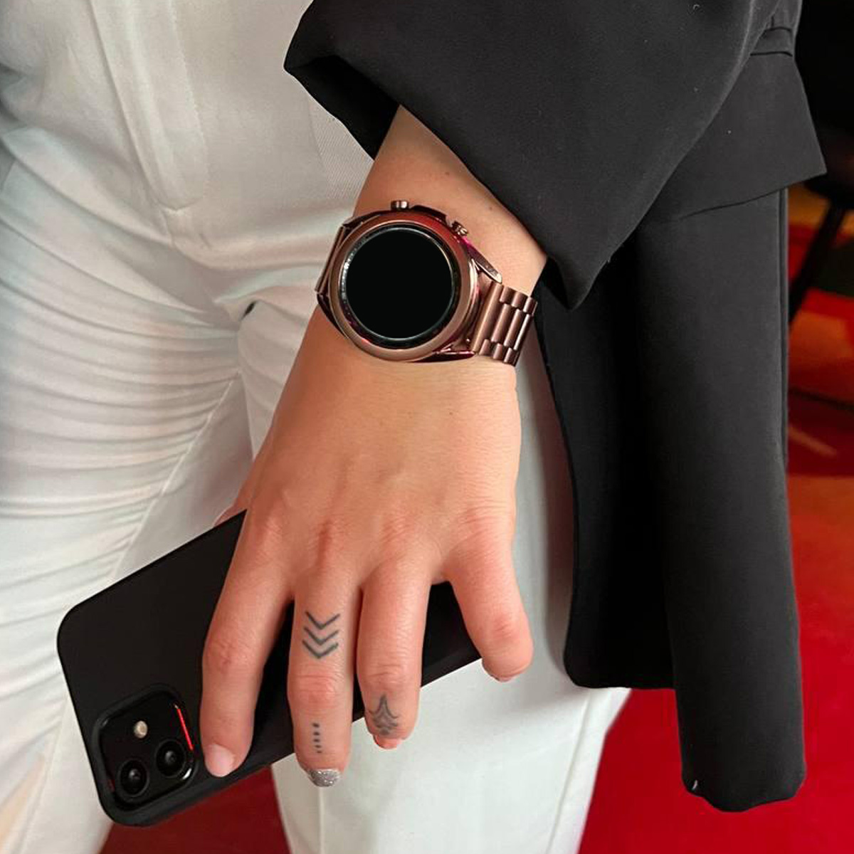 Hand einer Person mit roségoldener Uhr und Telefon. Die Person trägt einen schwarzen Blazer und eine weiße Hose.