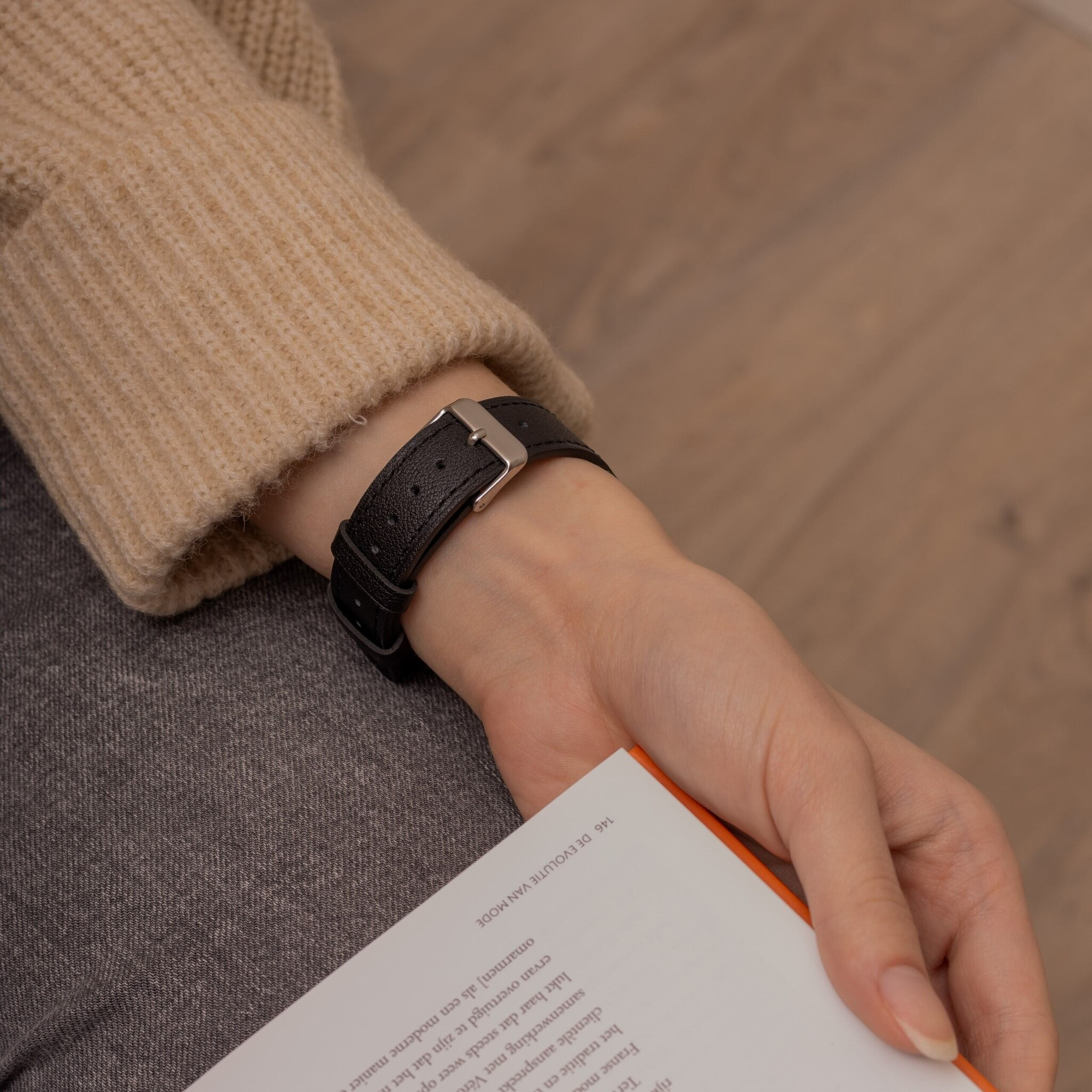 Eine Person, die eine schwarze Lederuhr mit silberner Schließe trägt und ein Buch hält.