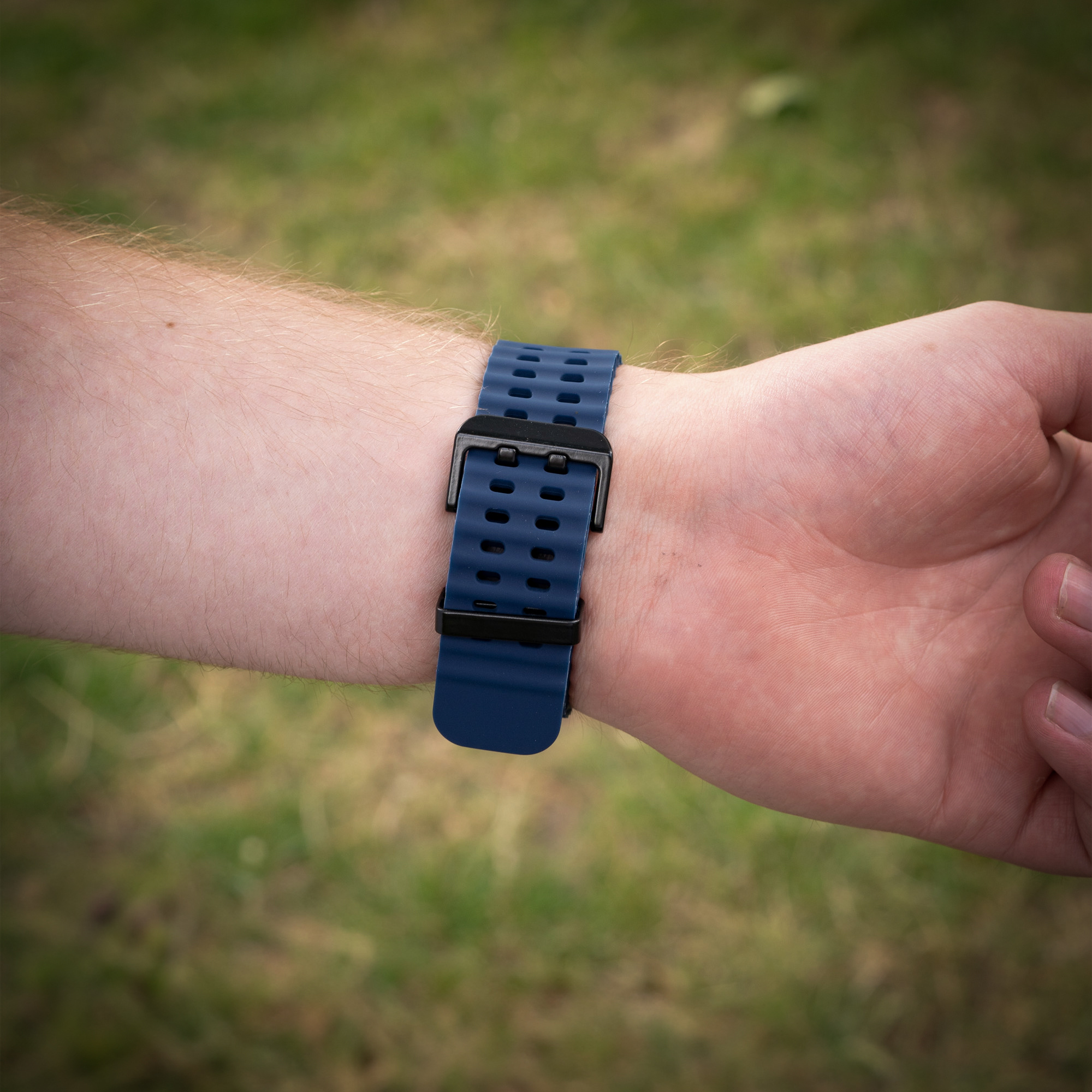 Ein dunkelblaues Uhrenarmband an einem Handgelenk mit schwarzer Schnalle, draußen im grünen Gras.