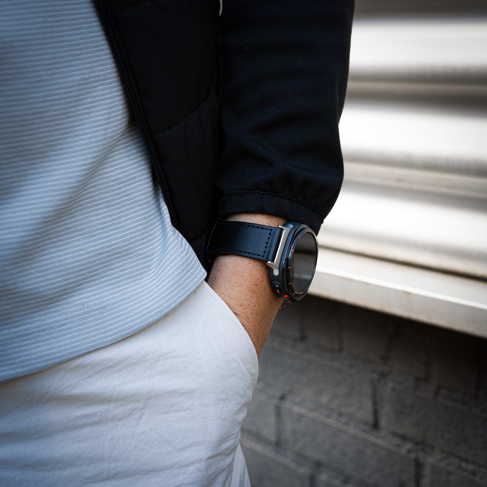Eine Person, die eine schwarze Uhr mit schwarzem Lederarmband trägt. Eine Ziegelmauer sichtbar.