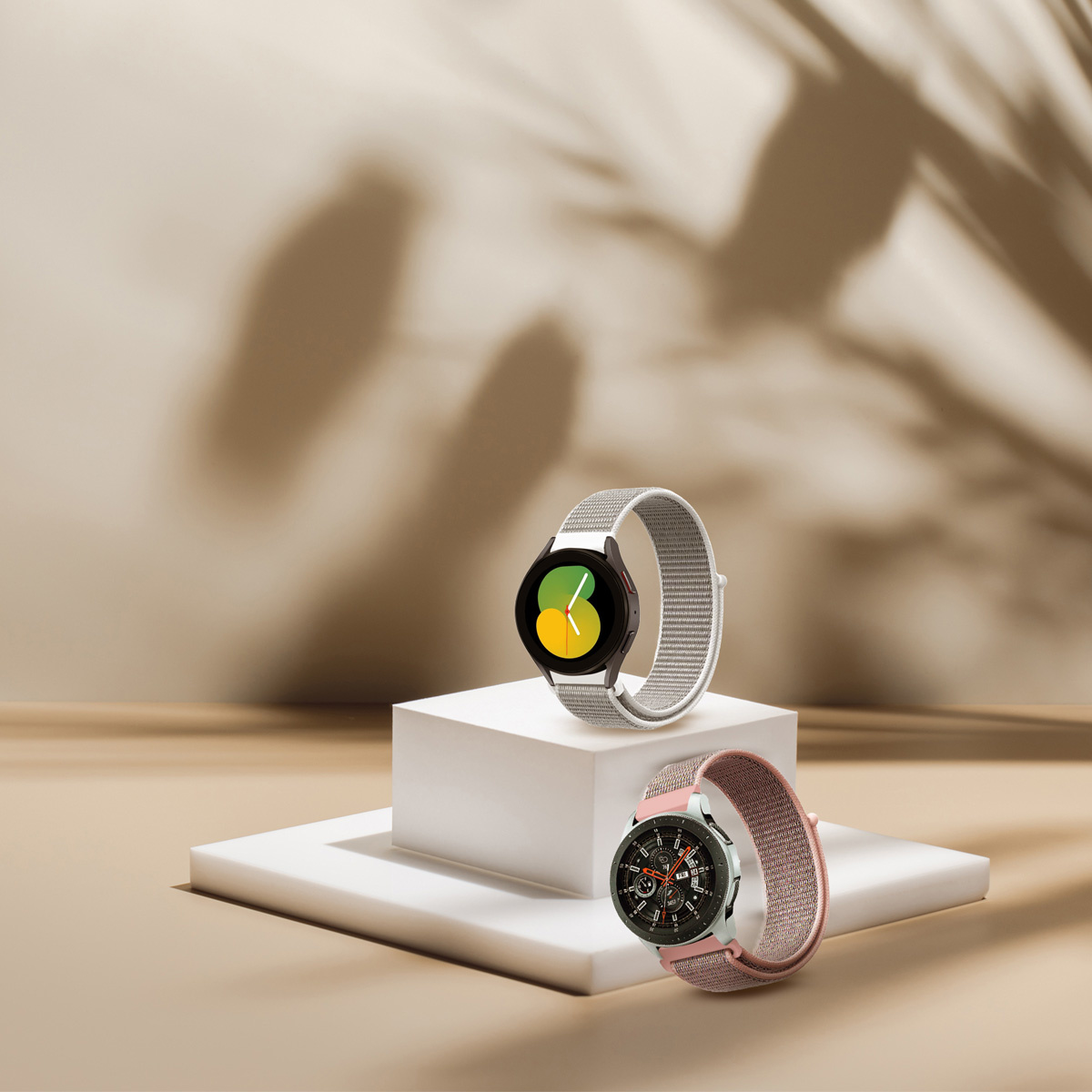 Zwei Smartwatches auf einem weißen Podest. Eine hat einen schwarzen Bildschirm, die andere ein rosa Armband.