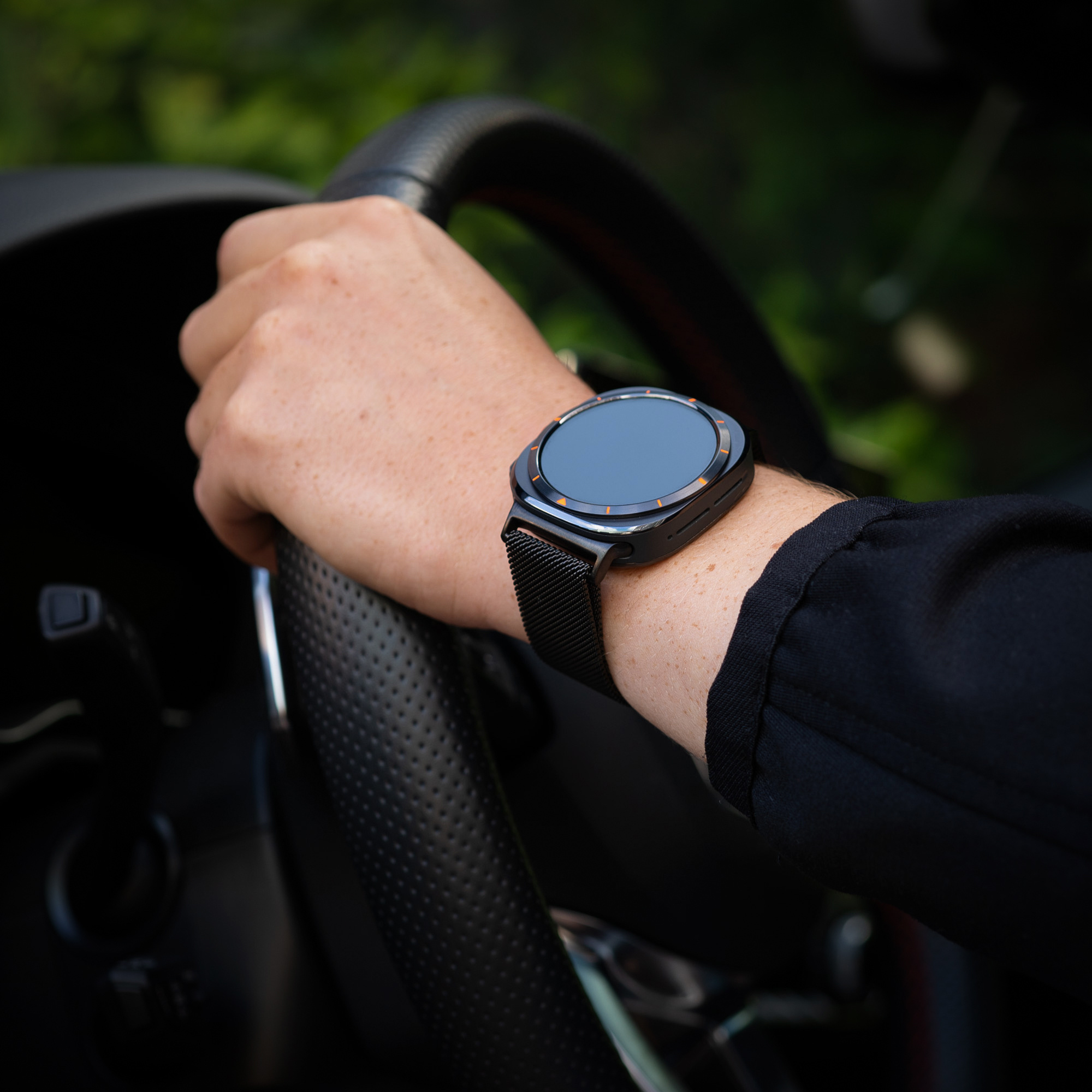 Hand mit schwarzer Smartwatch. Lenkrad und schwarze Jacke im Hintergrund sichtbar.