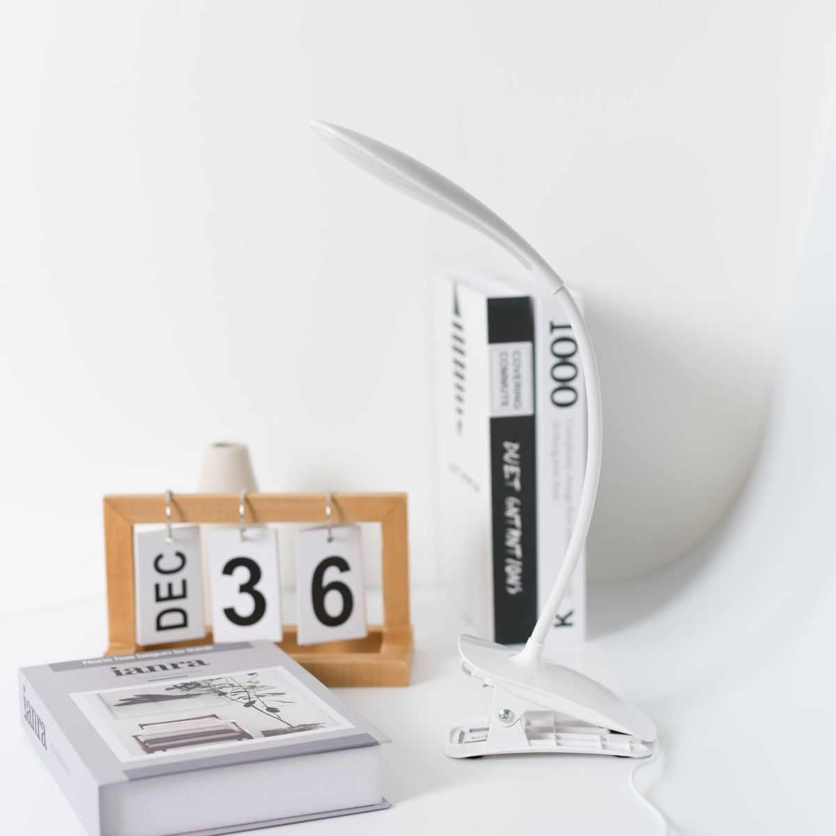 Lámpara de escritorio blanca sobre un escritorio blanco. El calendario muestra DIC 36. Libro y jarrón pequeño.
