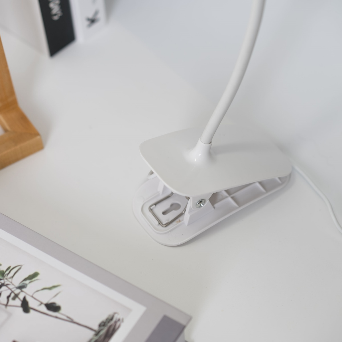 Pinza de lámpara de escritorio blanca. Está sobre una mesa blanca. La escena tiene un marco y libros.