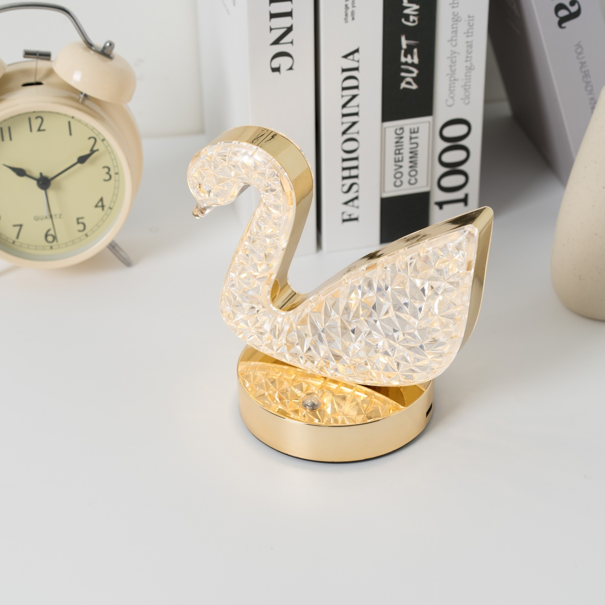 Una lámpara decorativa dorada en forma de cisne. Está sobre una superficie blanca con libros y un despertador al fondo.