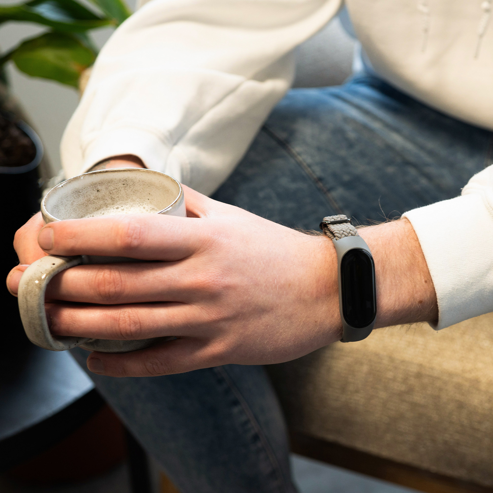 Person hält eine Tasse, trägt eine graue Smartwatch. Die Person trägt einen weißen Sweatshirt.