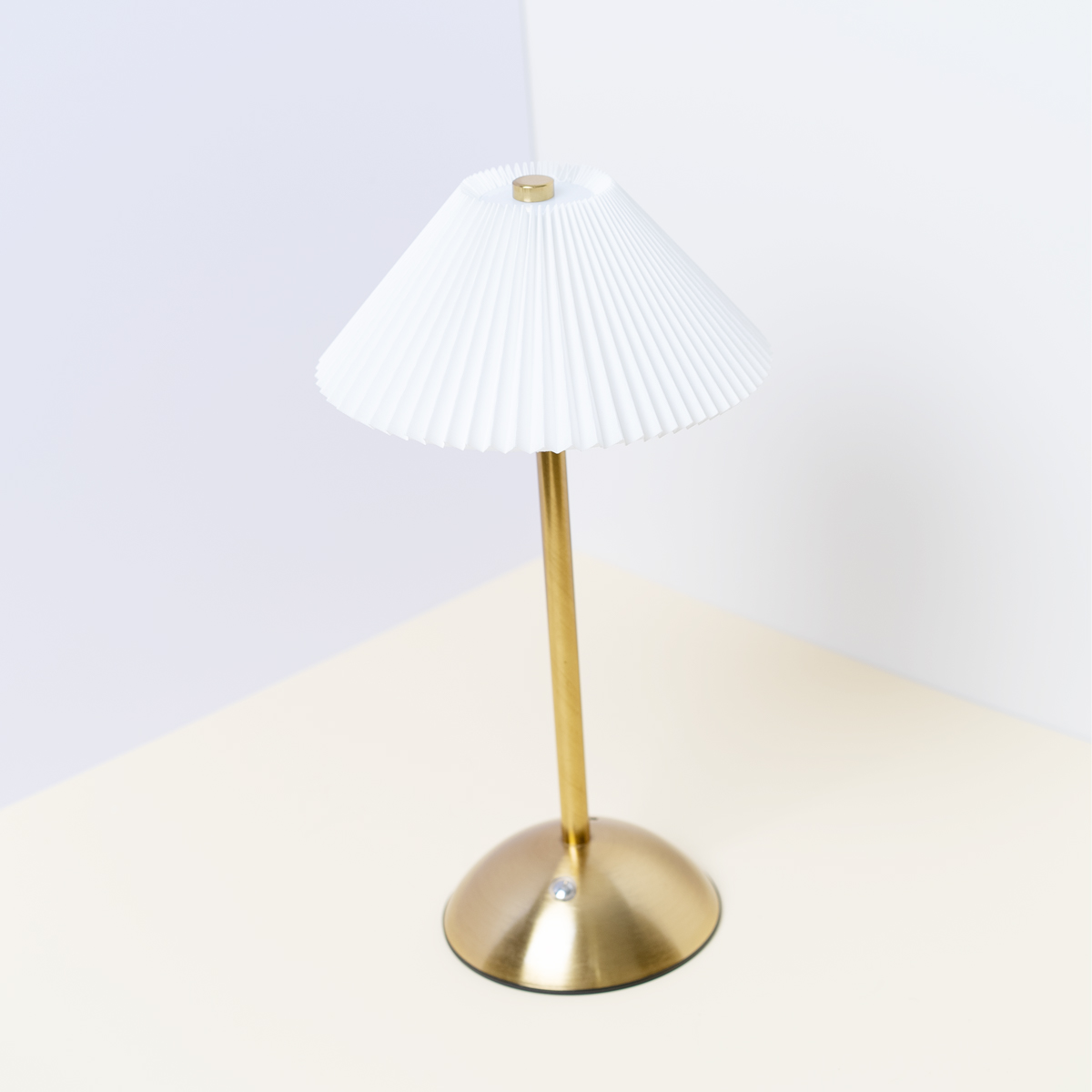 Lámpara de mesa dorada con pantalla plisada blanca, colocada sobre una superficie de dos tonos.