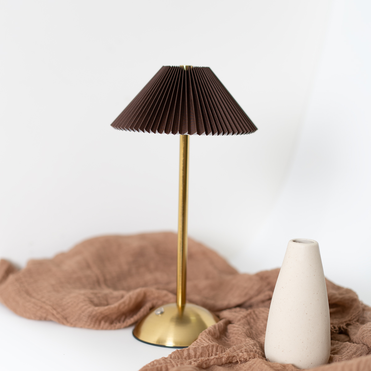 Una pequeña lámpara de mesa con una pantalla plisada marrón y un soporte dorado. Hay un jarrón beige al lado.