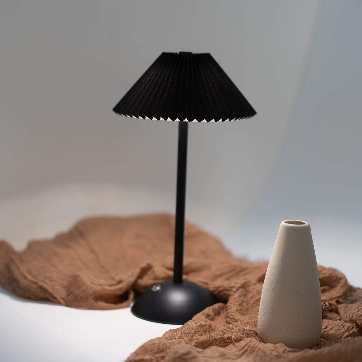 Lámpara de mesa negra con pantalla plisada. Un jarrón blanco y una tela marrón están cerca. Fondo blanco.