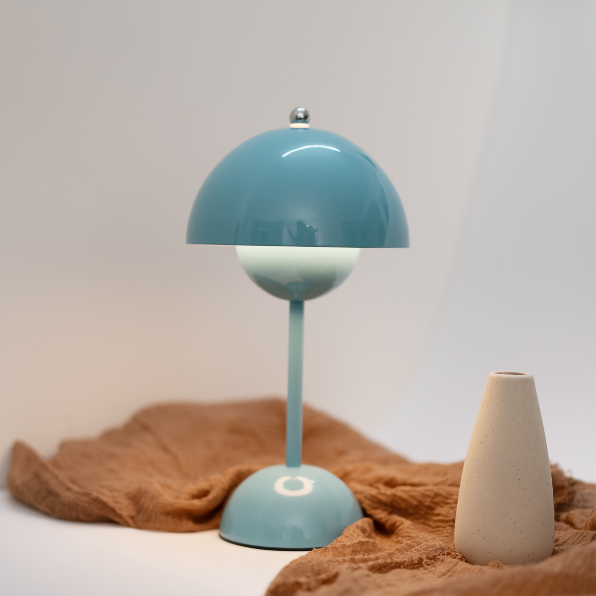 Lámpara de mesa azul con parte superior y base curvas, y un jarrón beige. Debajo hay una tela marrón.