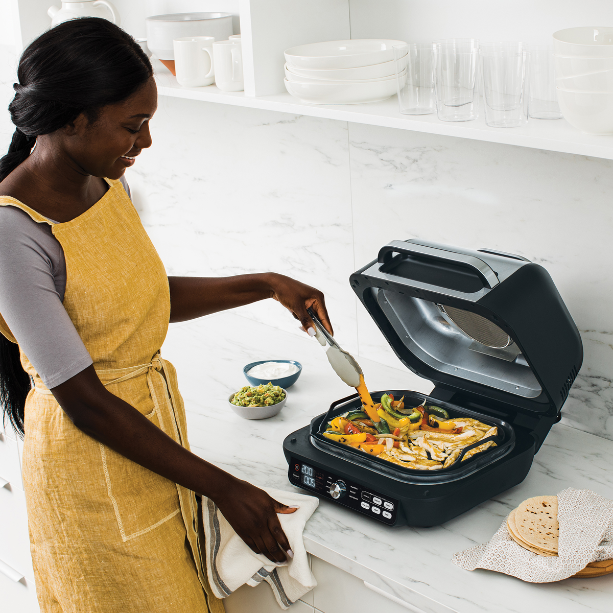 Eine Frau grillt Essen mit einem Ninja Woodfire Grill. Zutaten, Tortillas und Beilagen stehen auf der Theke.