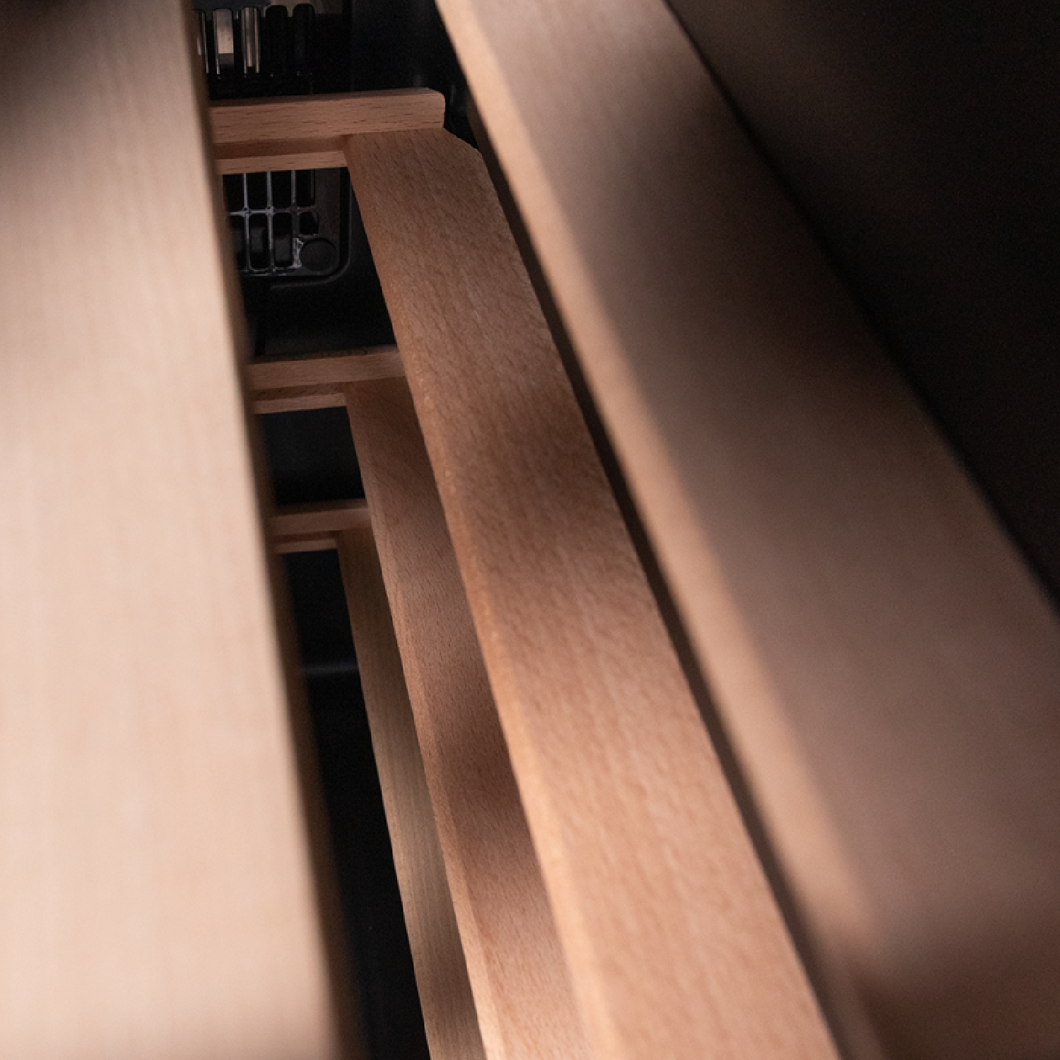 Wooden shelves inside a cabinet, viewed from an angle. Light brown wood contrasts with a dark interior. A metal grill is visible.