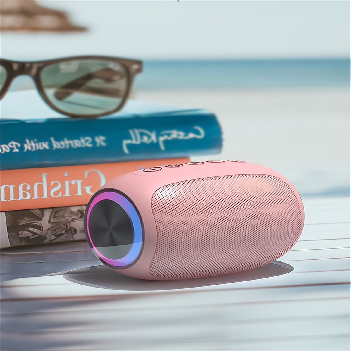 Rosa Lautsprecher mit Bedienelementen neben Büchern und Sonnenbrille am Strand.
